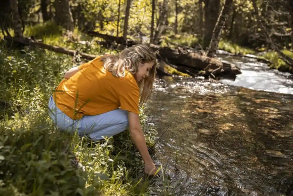 Mulher jovem de cabelos claros e camiseta laranja colocando a mão em agua cristalina de um rio em uma floresta fechada durante o dia em uma área de preservação permanente.